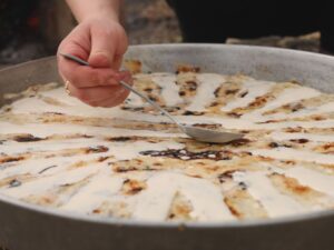Fli being prepared by hand in a large round pan, with cream being spooned over the layered crepes, a traditional dish from northern Albania